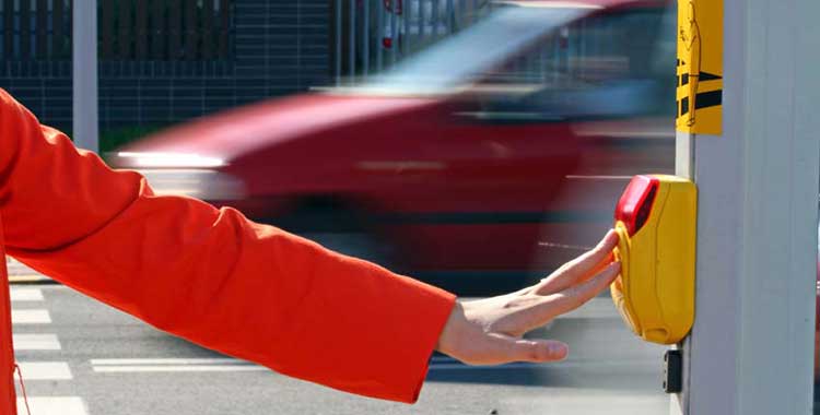 A pedestrian pushes the crosswalk button, but speeding cars nearby show risk of a pedestrian accident.